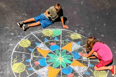 A boy and a girl draw a large chalk mandala on a sidewalk